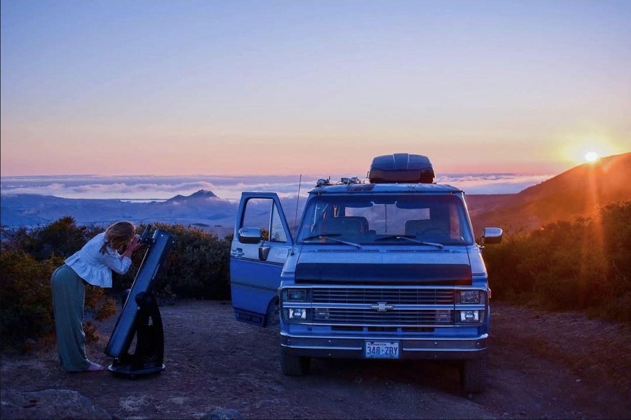 A person looking through a large telescope next to a parked project van above a fog layer at sunset