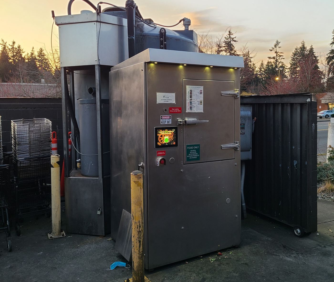 A WISErg Harvestor installed behind a grocery store at dusk, showing the stainless enclosure, control panel, and digester tanks
