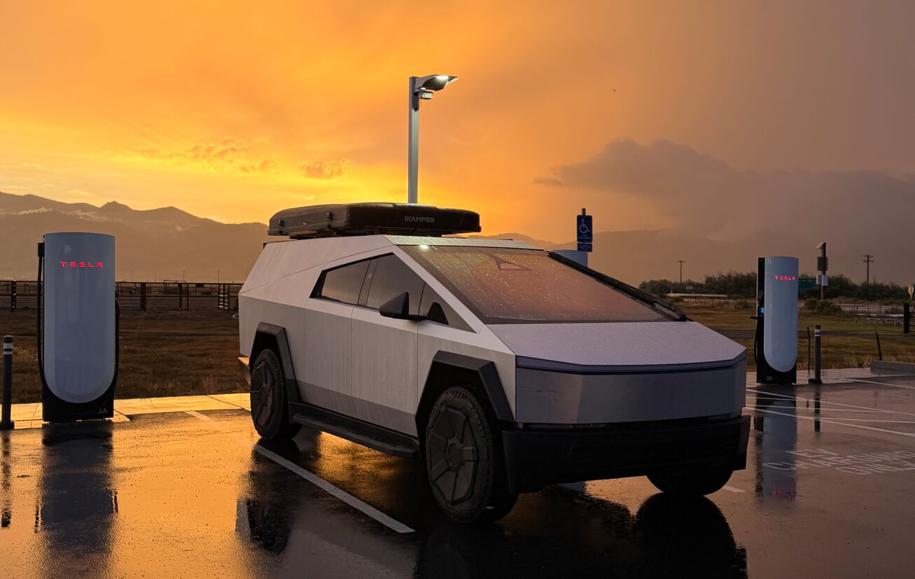 A stainless Cybertruck with the Space Campers cap and a rooftop tent, parked at a Tesla Supercharger at sunset
