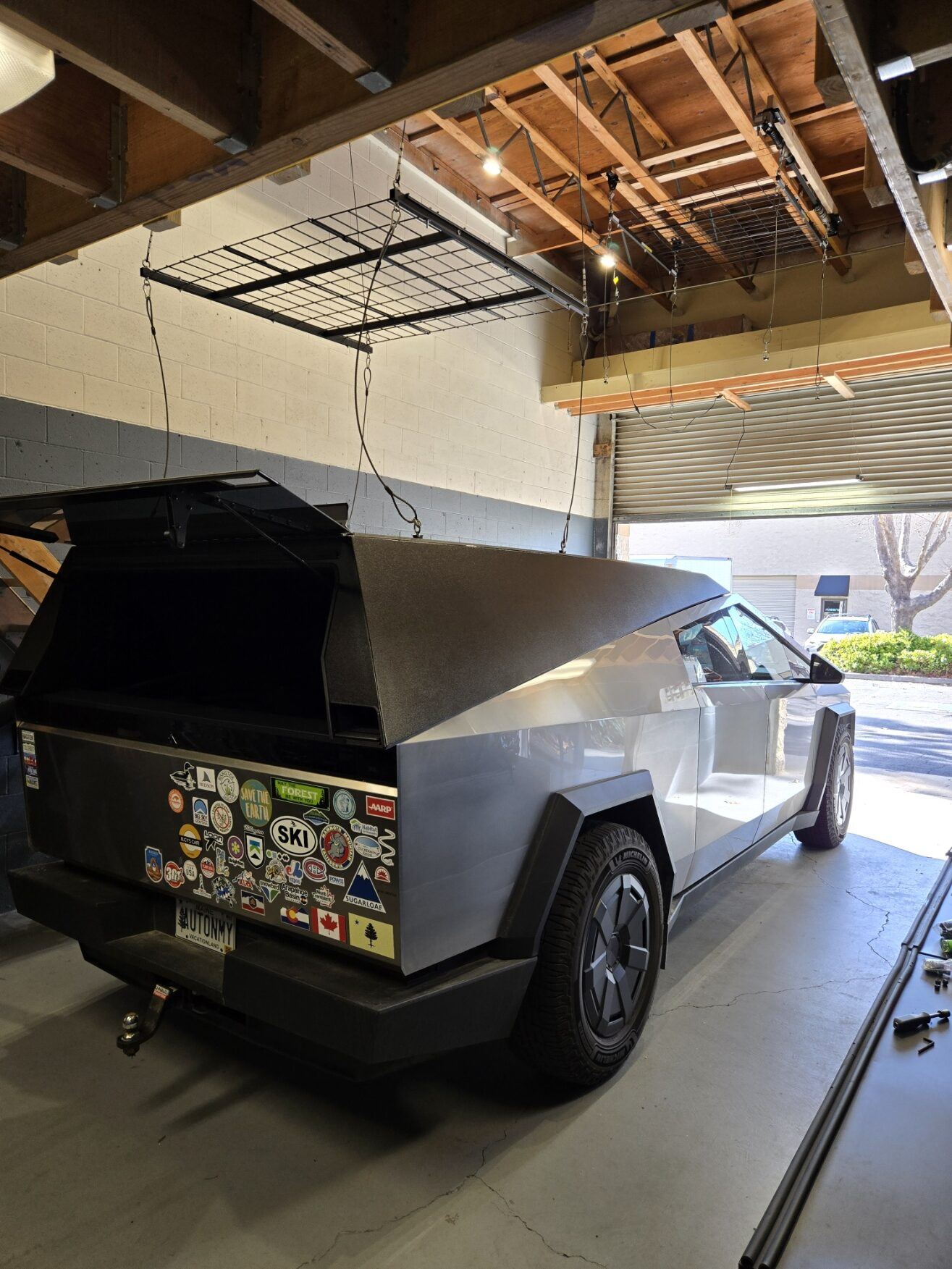 A stainless Cybertruck with the rear cap hatch open, sticker-covered tailgate, parked in the shop