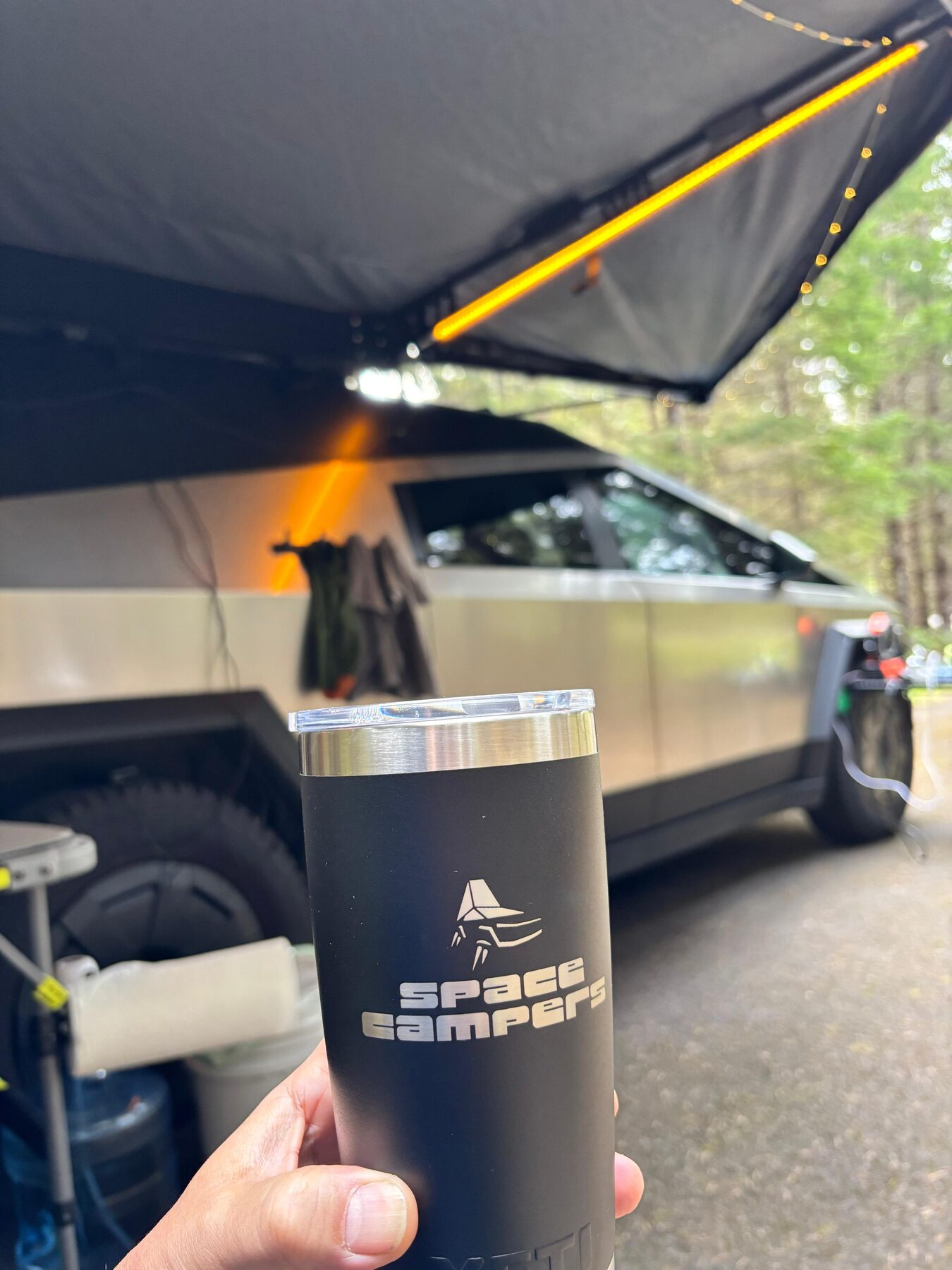 A Space Campers tumbler held up in front of the truck with the awning and string lights out