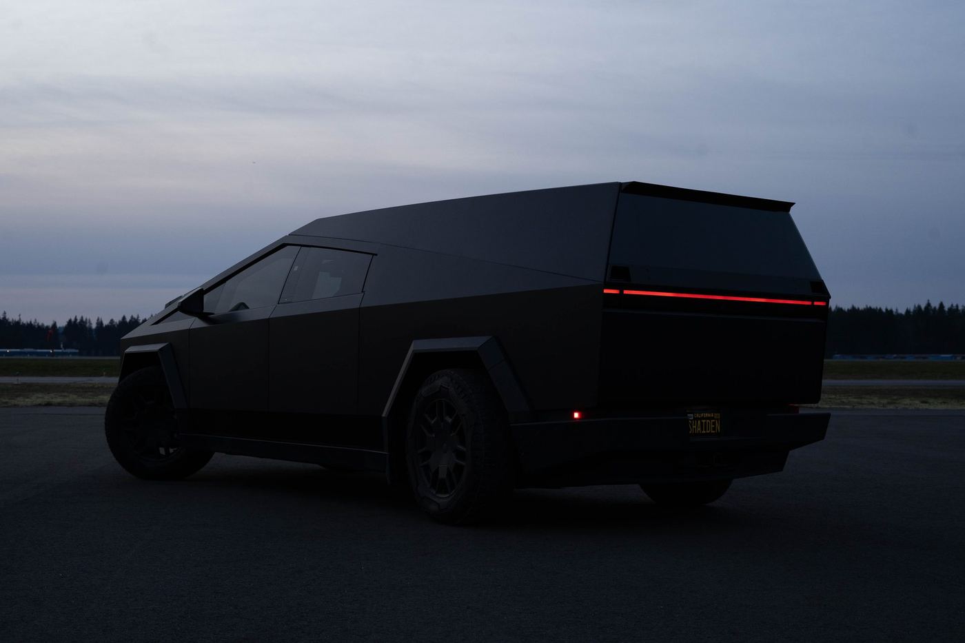 A matte black Cybertruck and color-matched cap parked on a runway at dusk, rear three-quarter view with the taillight bar lit