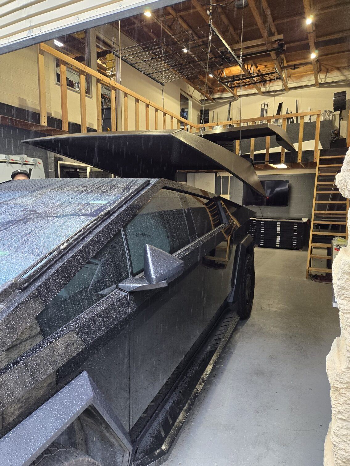 A black Cybertruck in the shop with both cap side hatches raised on their gas struts