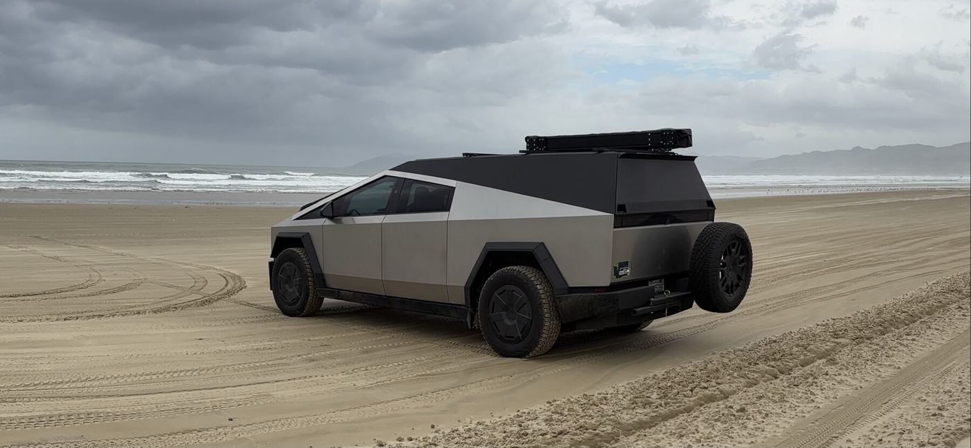 A Cybertruck with the cap and a roof-mounted tent box driving on wet sand at the beach