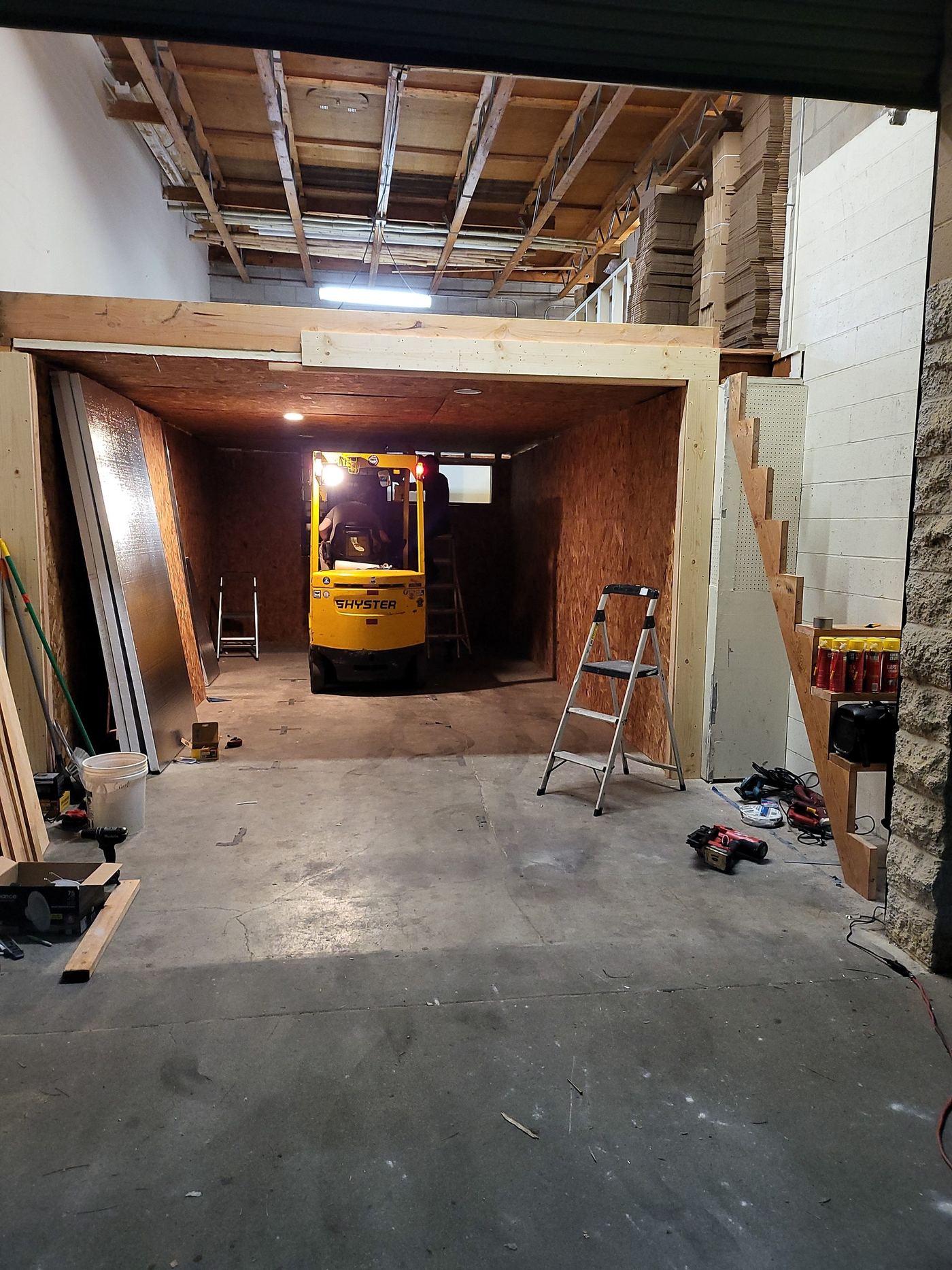 A small yellow forklift backed into the framed cold room during construction, insulation panels leaning against the wall