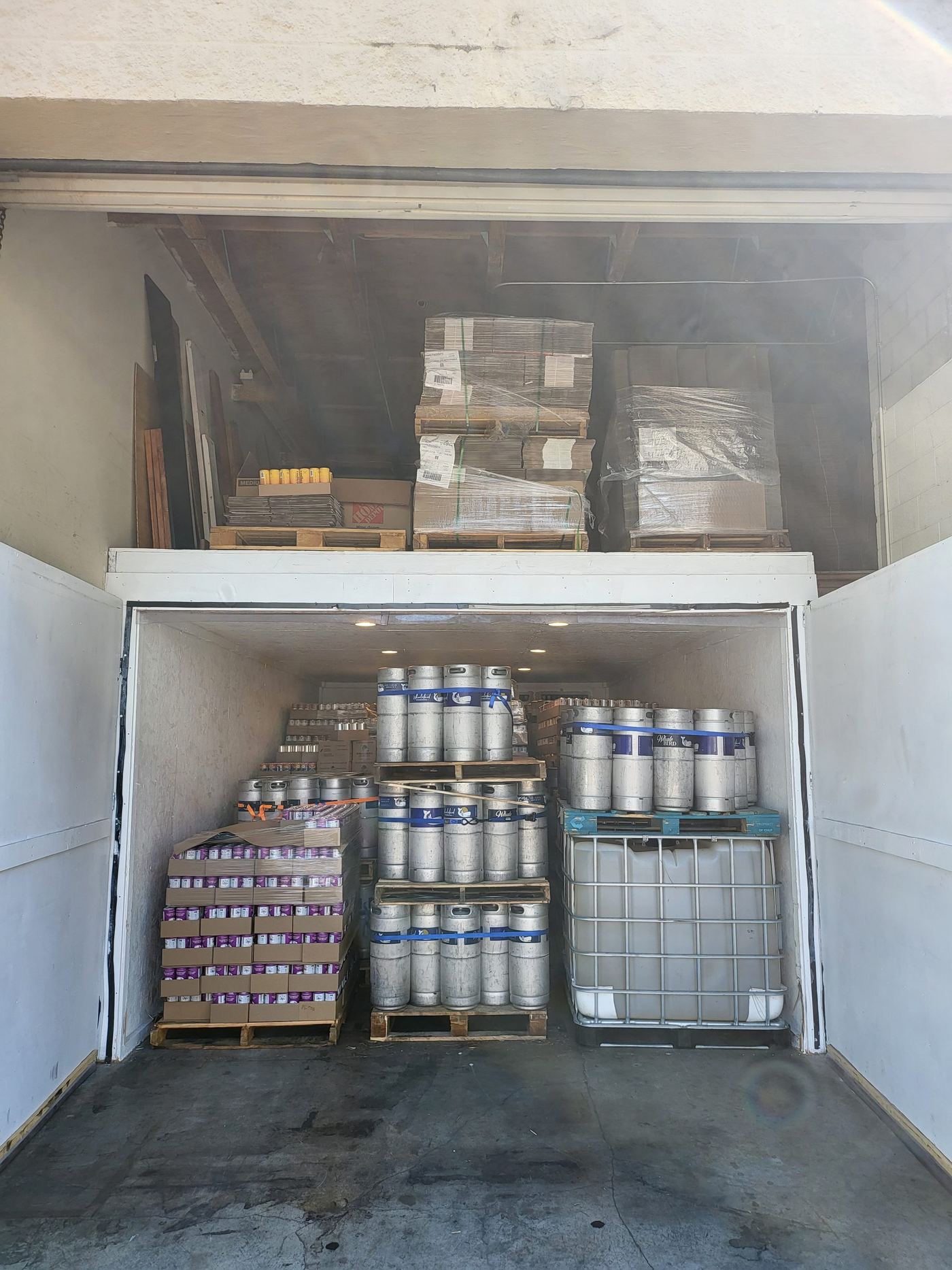 Kegs and a plastic tote packed into the finished cold room, seen through a hazy doorway past shelves of boxed stock