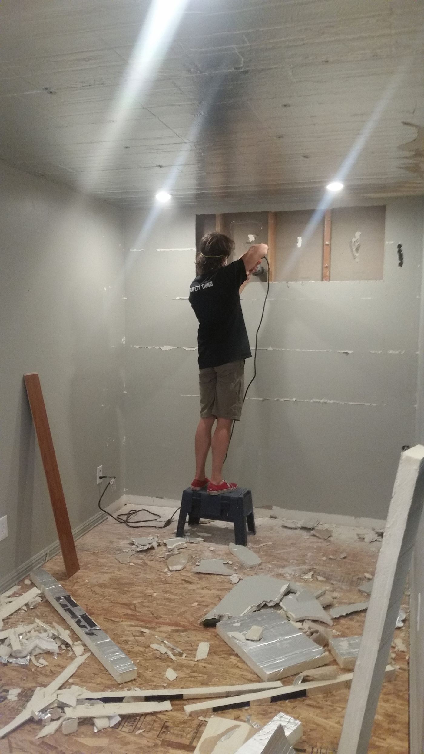 Standing on a step stool to cut an opening in the wall for the through-wall AC units, drywall scraps piled on the plywood floor