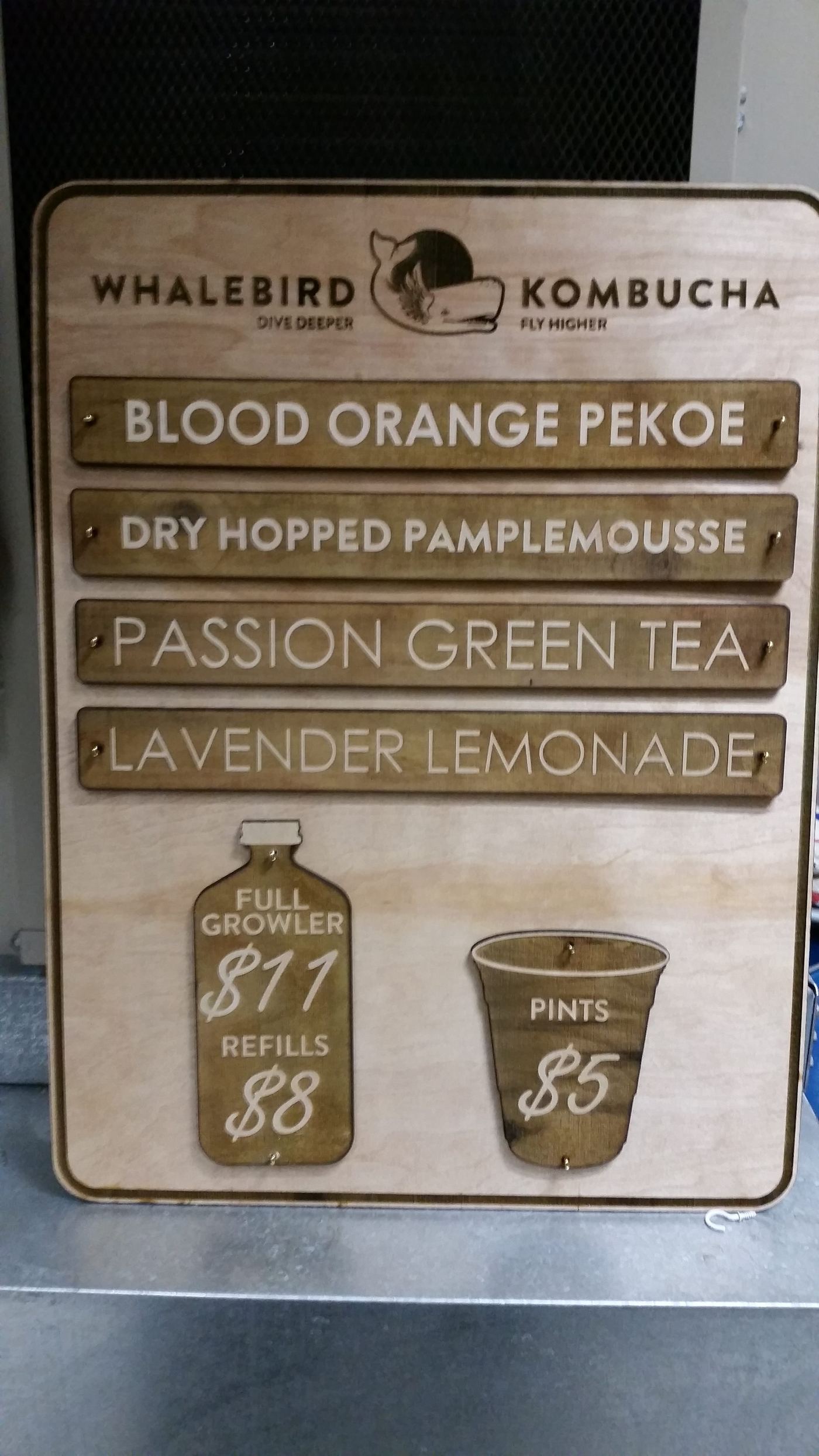 A wood flavor menu board listing four kombucha flavors alongside pint and growler pricing