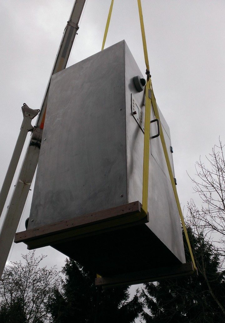 The stainless control enclosure mid-lift by crane against an overcast sky, ready to be set at the install site